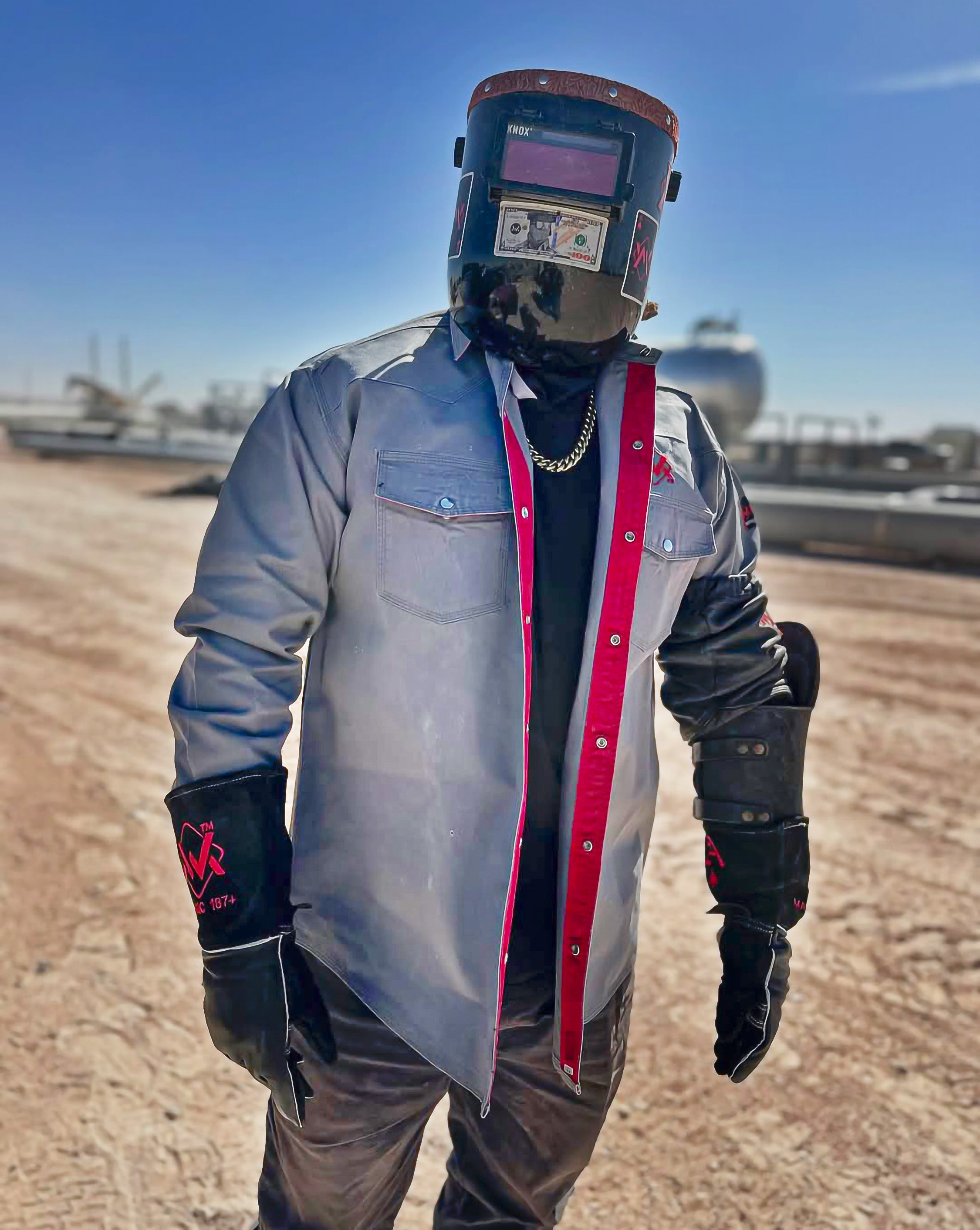 Person wearing a welding helmet, jacket, and gloves in an industrial setting.