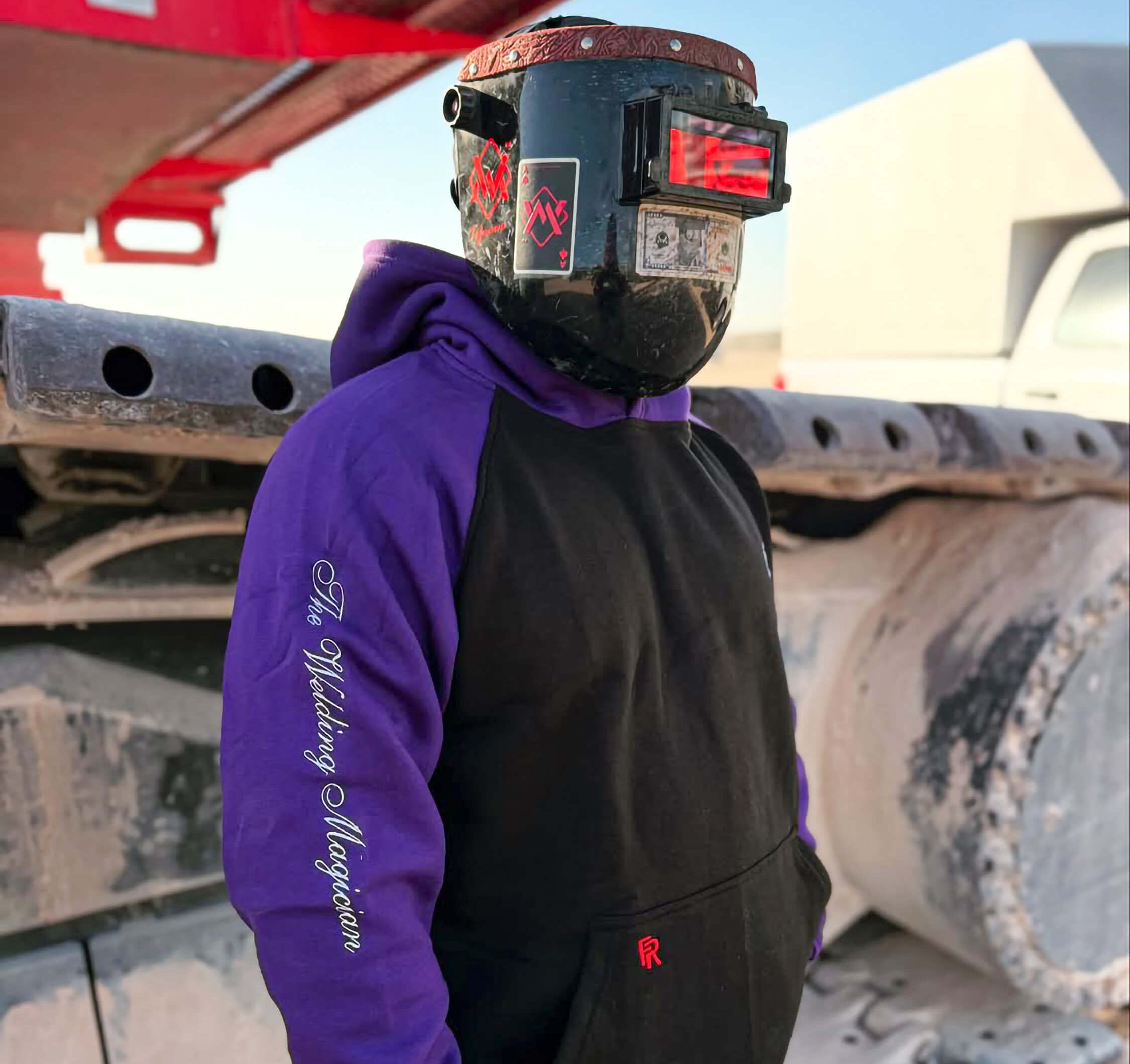 Person wearing a welding helmet and purple hoodie with text, standing next to large cylindrical objects.