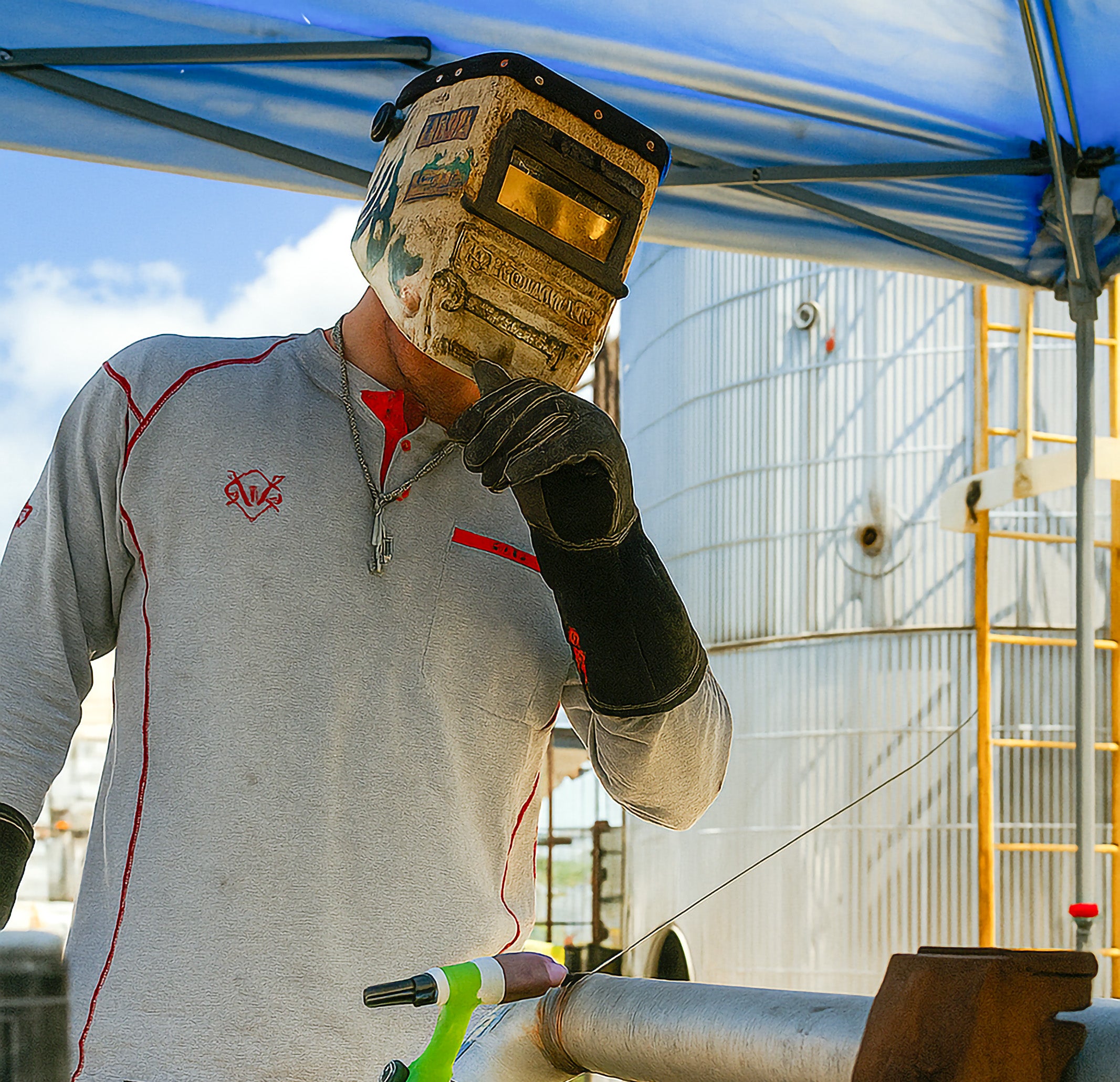 Person wearing a welding helmet and gloves, working on a metal pipe outdoors.