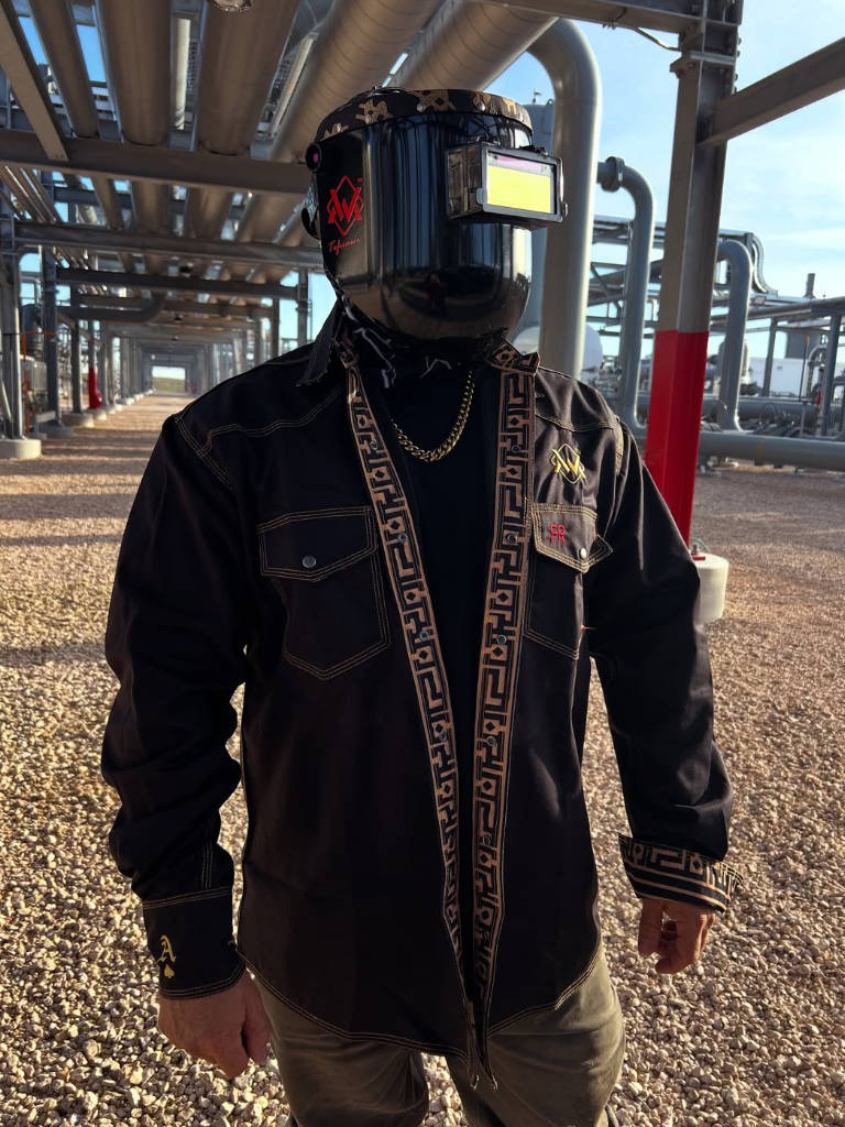 Person wearing a black jacket with a patterned strap and a helmet with a visor in an industrial setting.