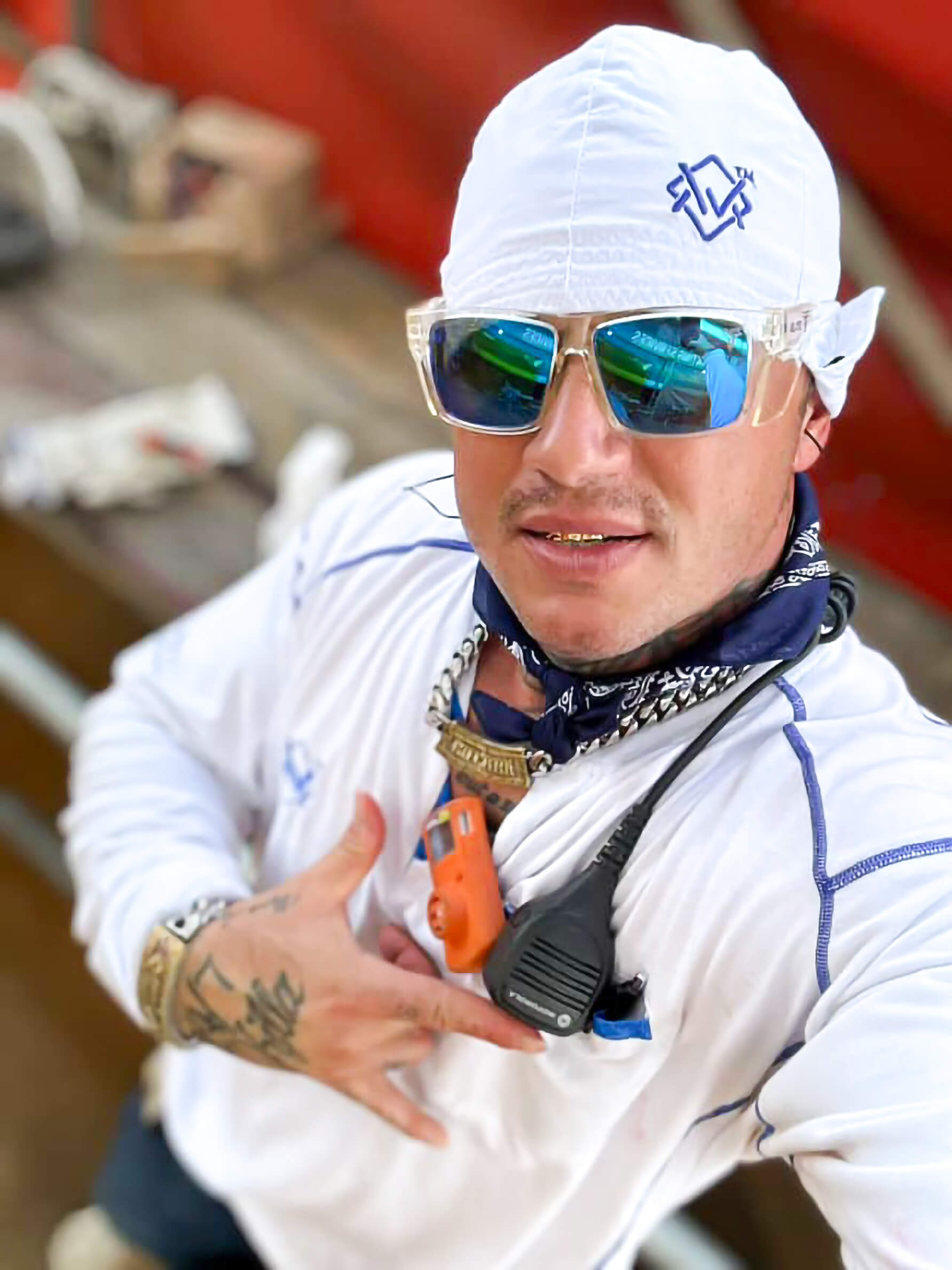 Person wearing a white beanie, sunglasses, and a white jacket with a bandana, sitting outdoors.