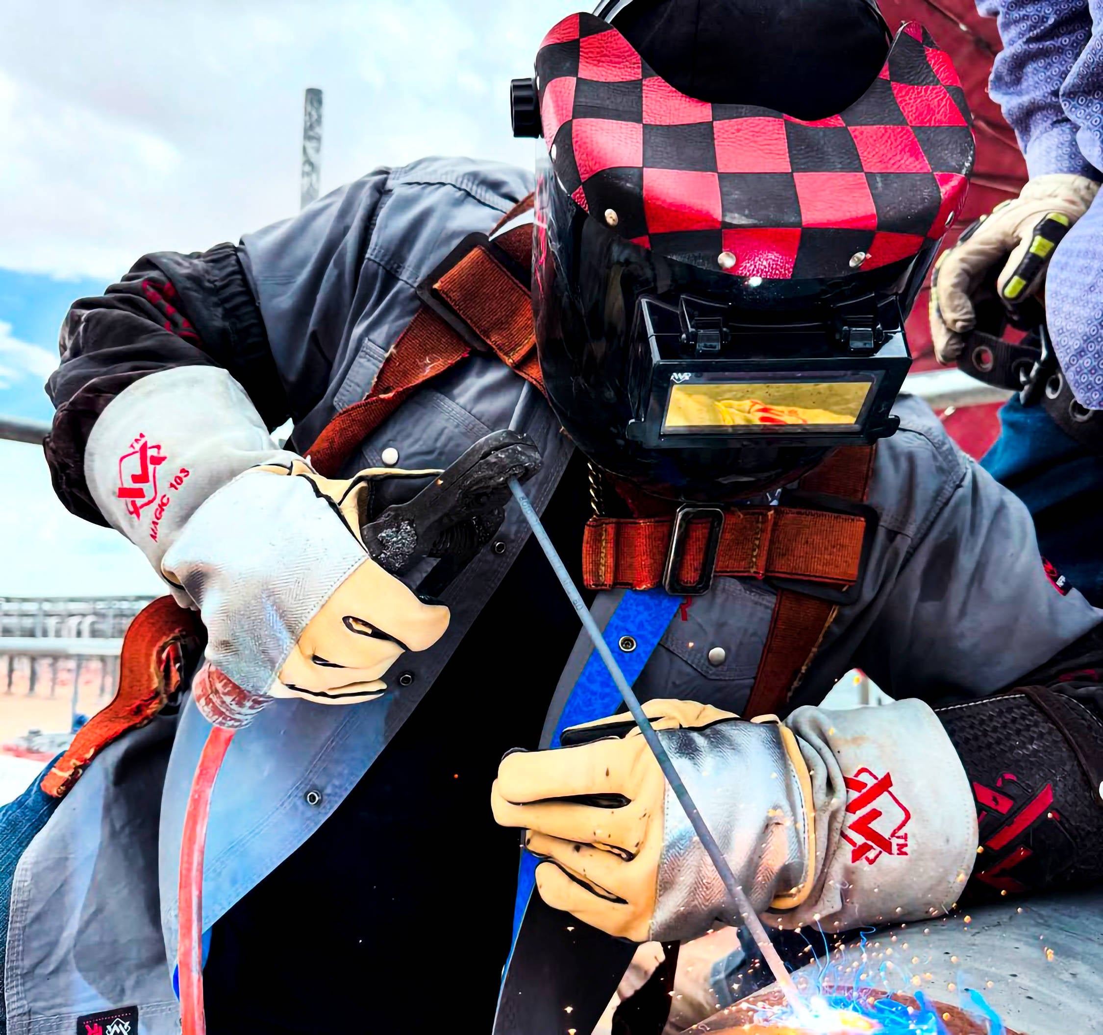 Welder in protective gear using a red-and-black welding hood outdoors.
