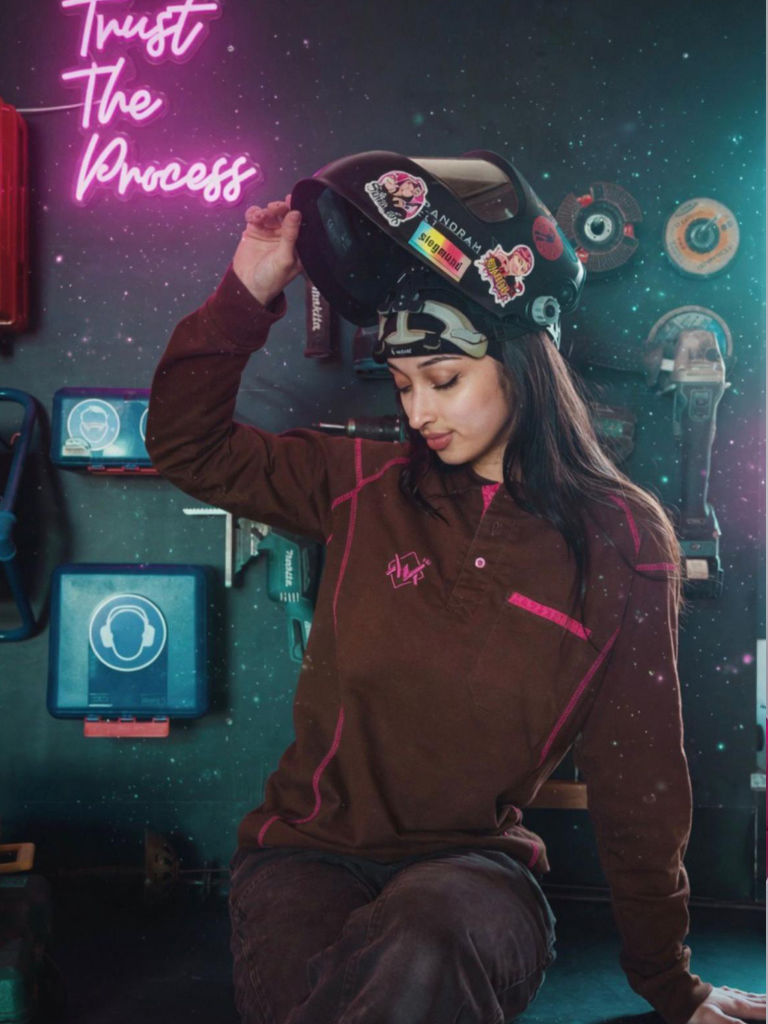A woman wearing a brown welding jacket and helmet sits in a workshop setting with a neon sign that reads 'Trust The Process' on the wall.