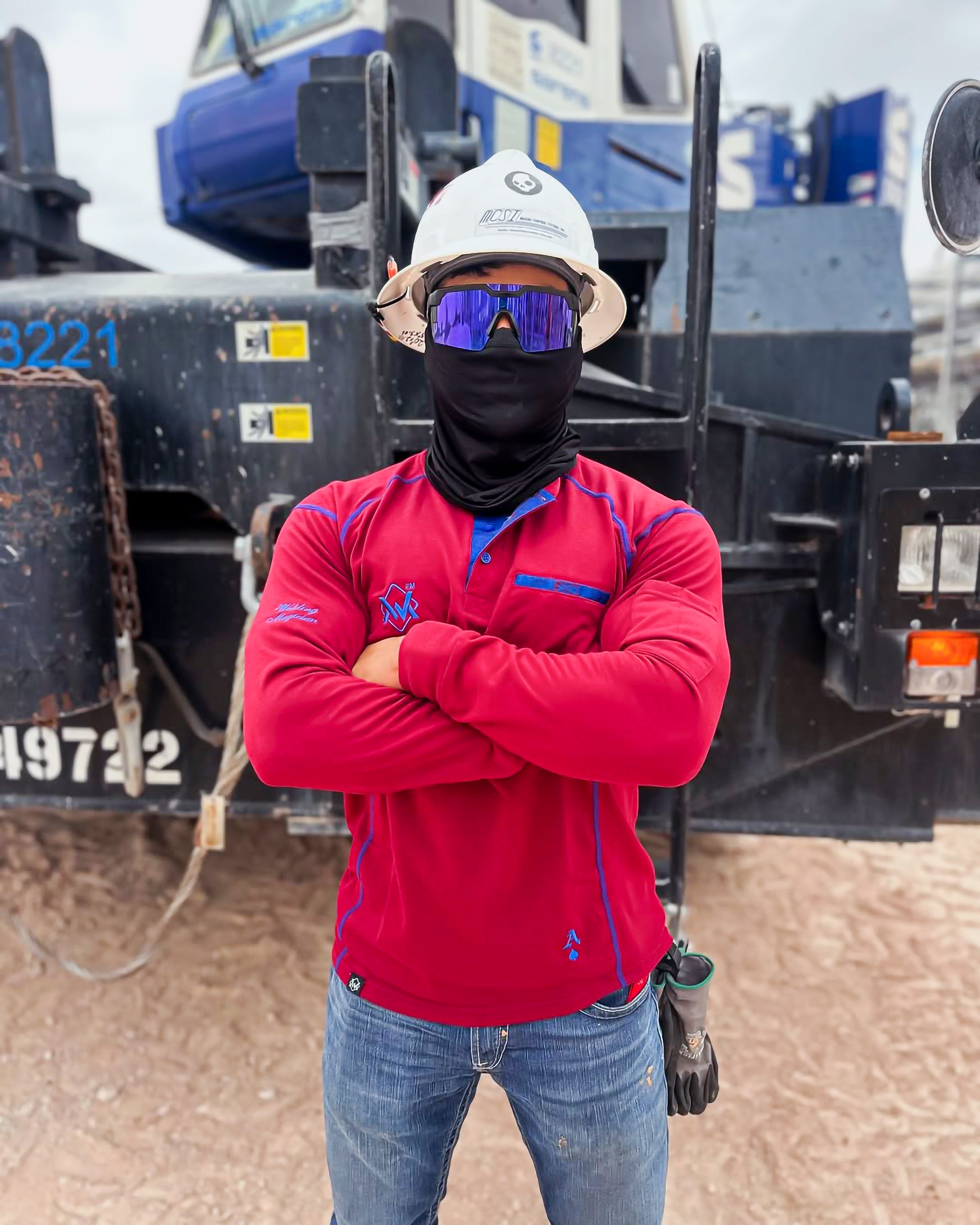 Person wearing a red shirt, white hard hat, and black mask standing in front of a large piece of machinery.