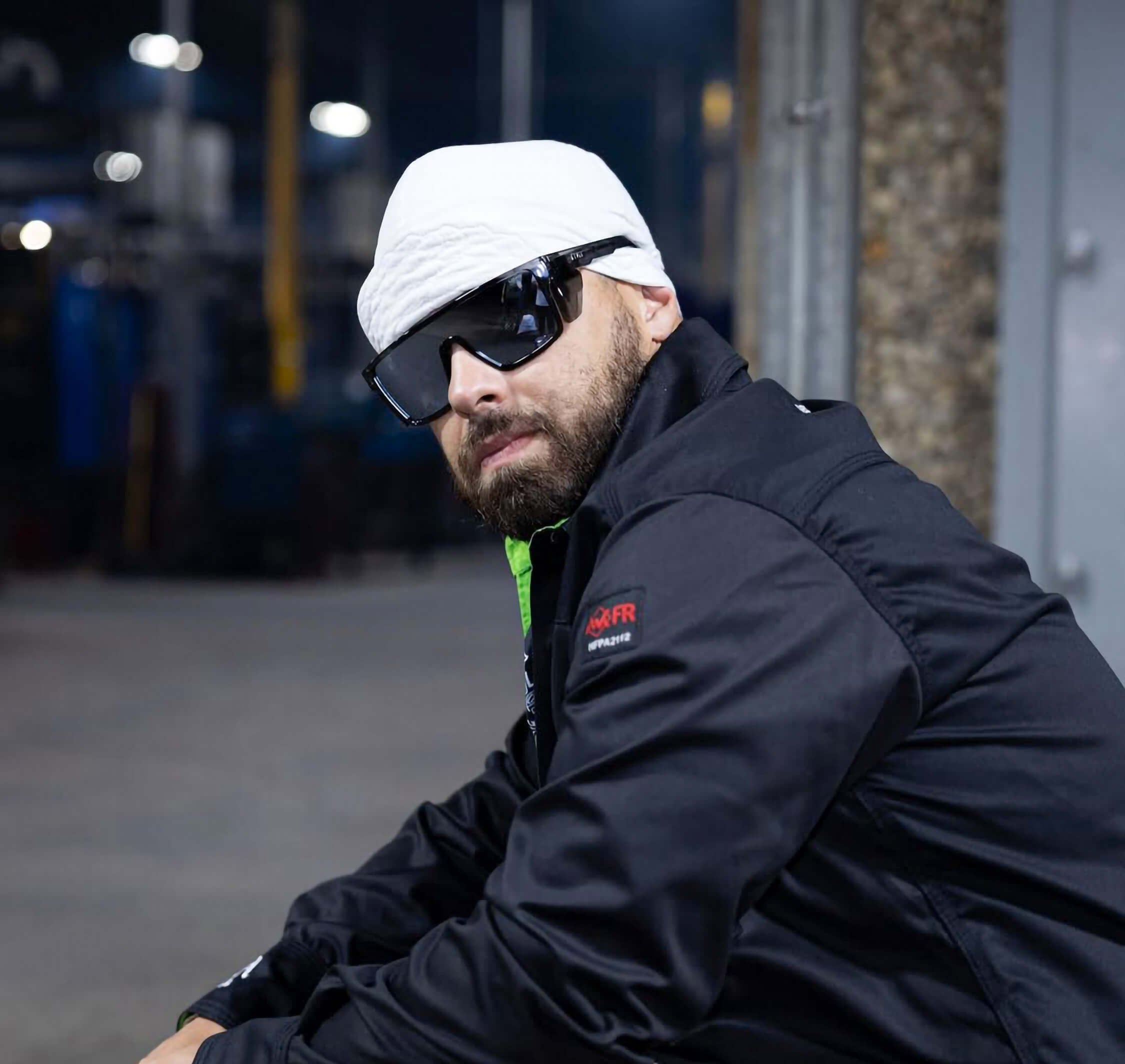 Man wearing a black jacket with a logo, white cap, and sunglasses in an urban setting.