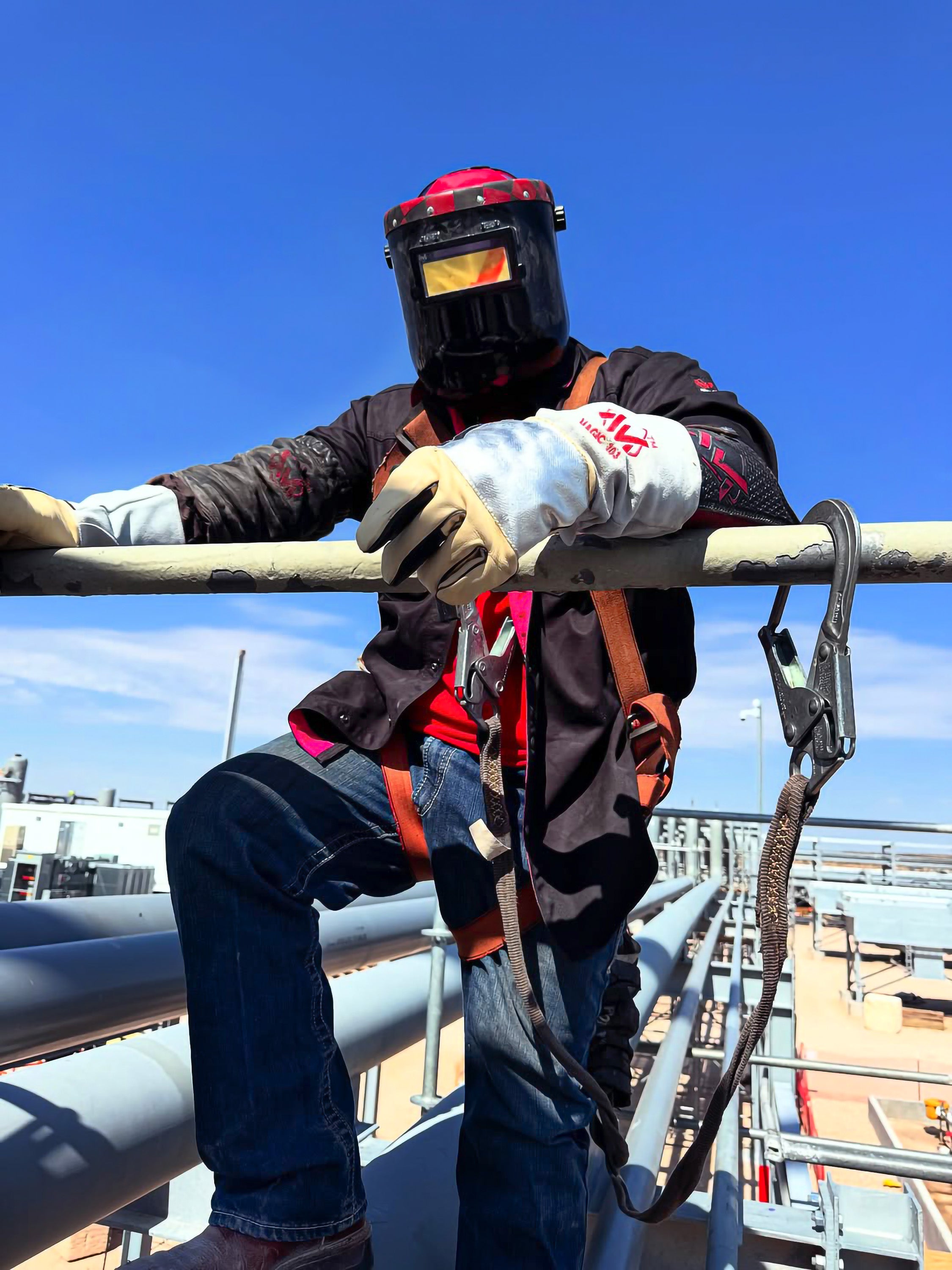 Person wearing a welding helmet and safety gear on a metal structure with a clear blue sky.