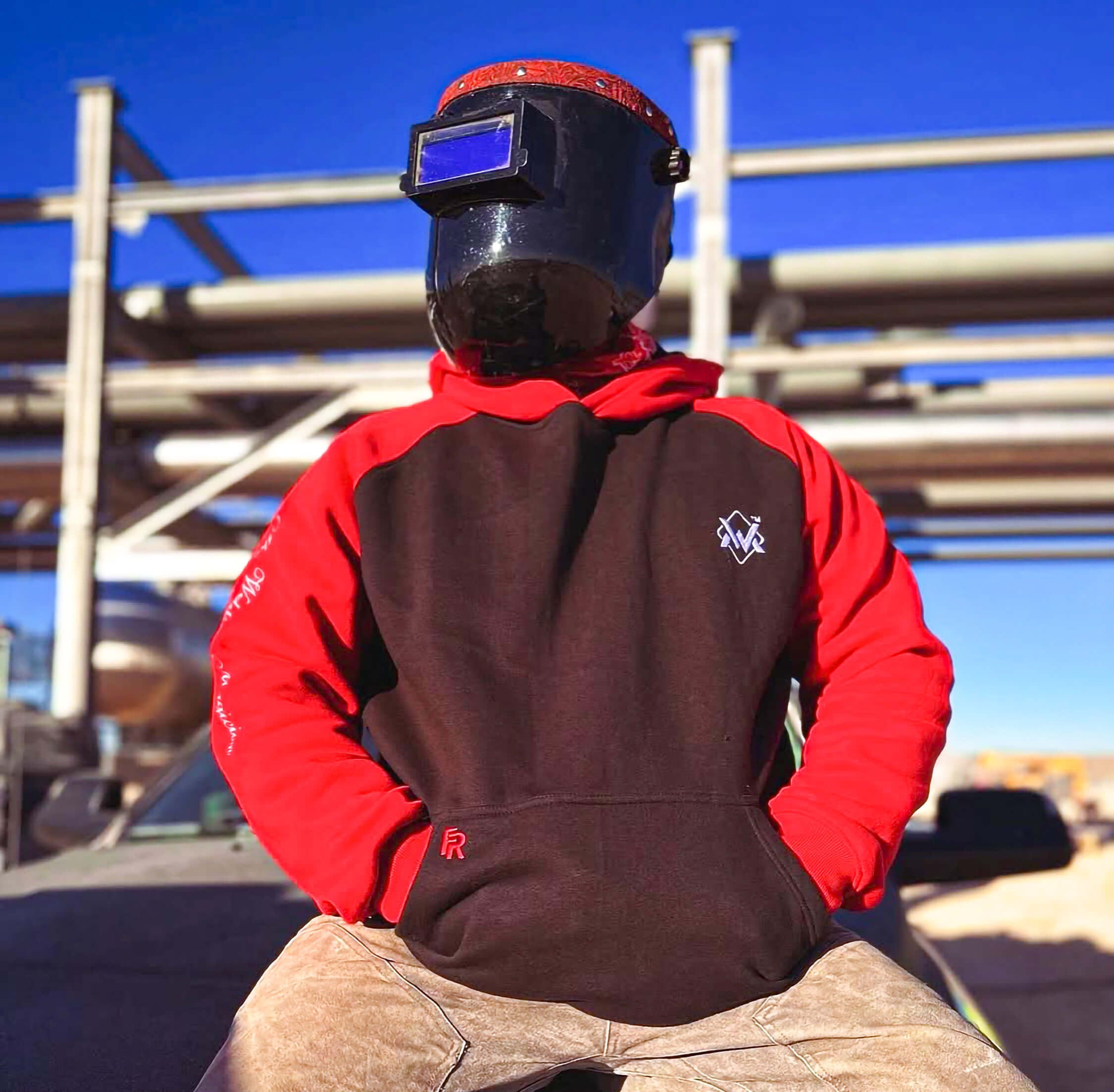 Person wearing a red and black welding helmet and protective clothing sitting on a car.
