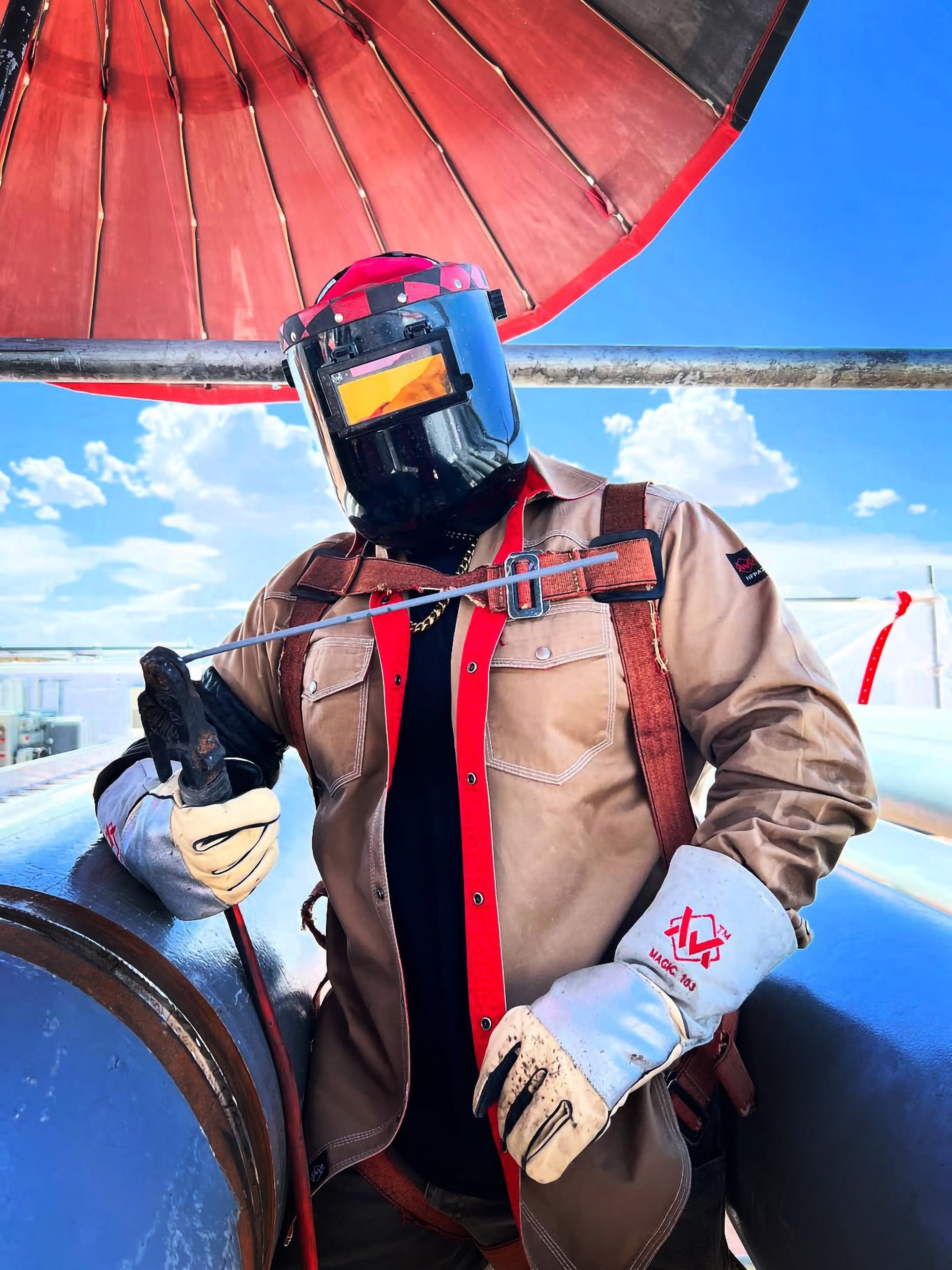 Person in a protective suit with a helmet and gloves, holding a hose against a blue sky with clouds.
