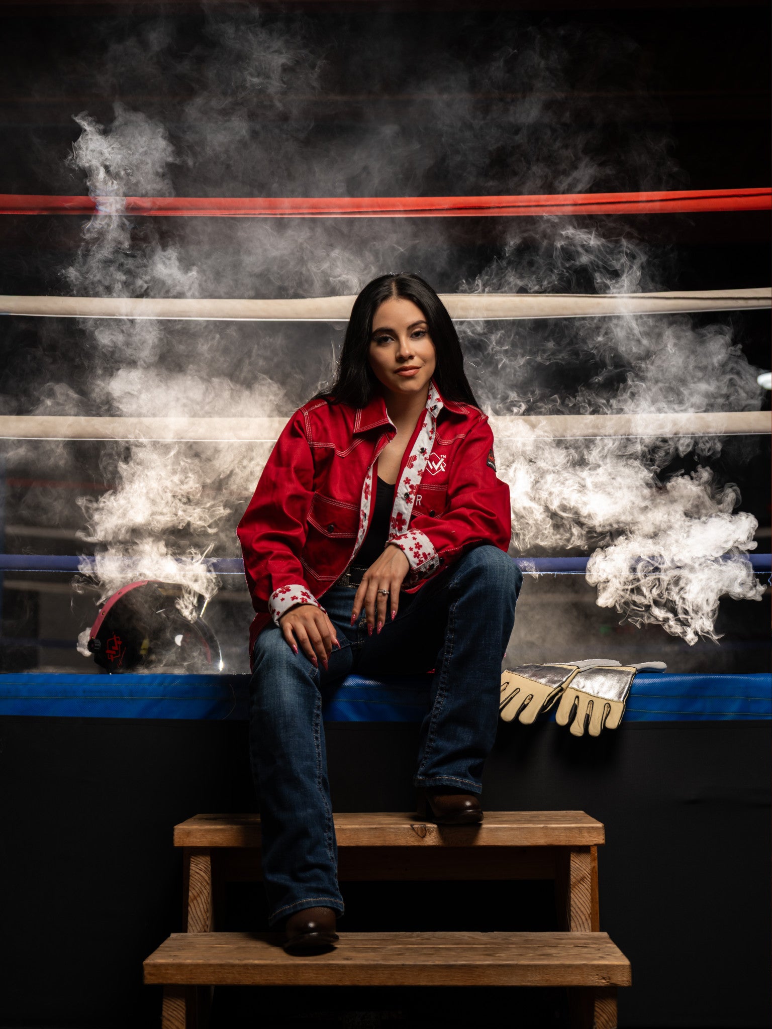 Person in a red jacket sitting on a wooden stool in a boxing ring with smoke effects.