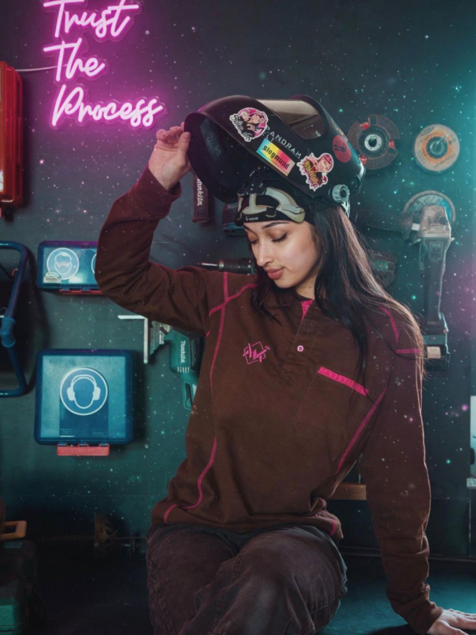 Person wearing a brown/pink long sleeve and helmet with stickers in a workshop setting with neon sign 'Trust The Process'.