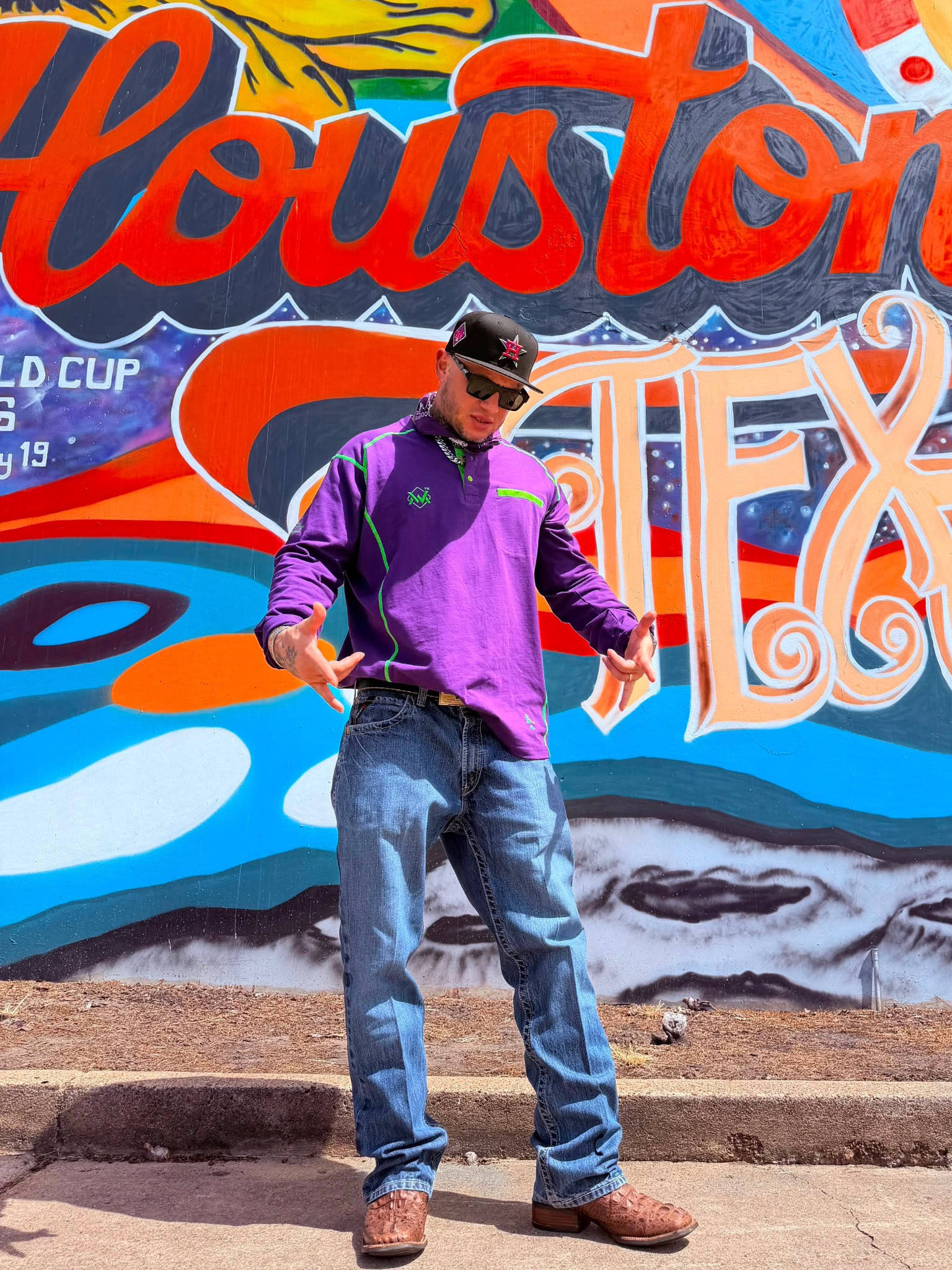 A man wearing a purple/joker long-sleeve quarter-zip pullover, blue jeans, brown boots, and a black cap stands in front of a colorful Houston, Texas graffiti mural.