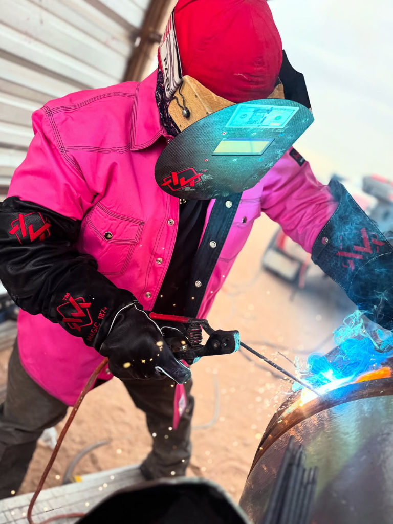 Person in pink jacket and red helmet welding metal with sparks flying