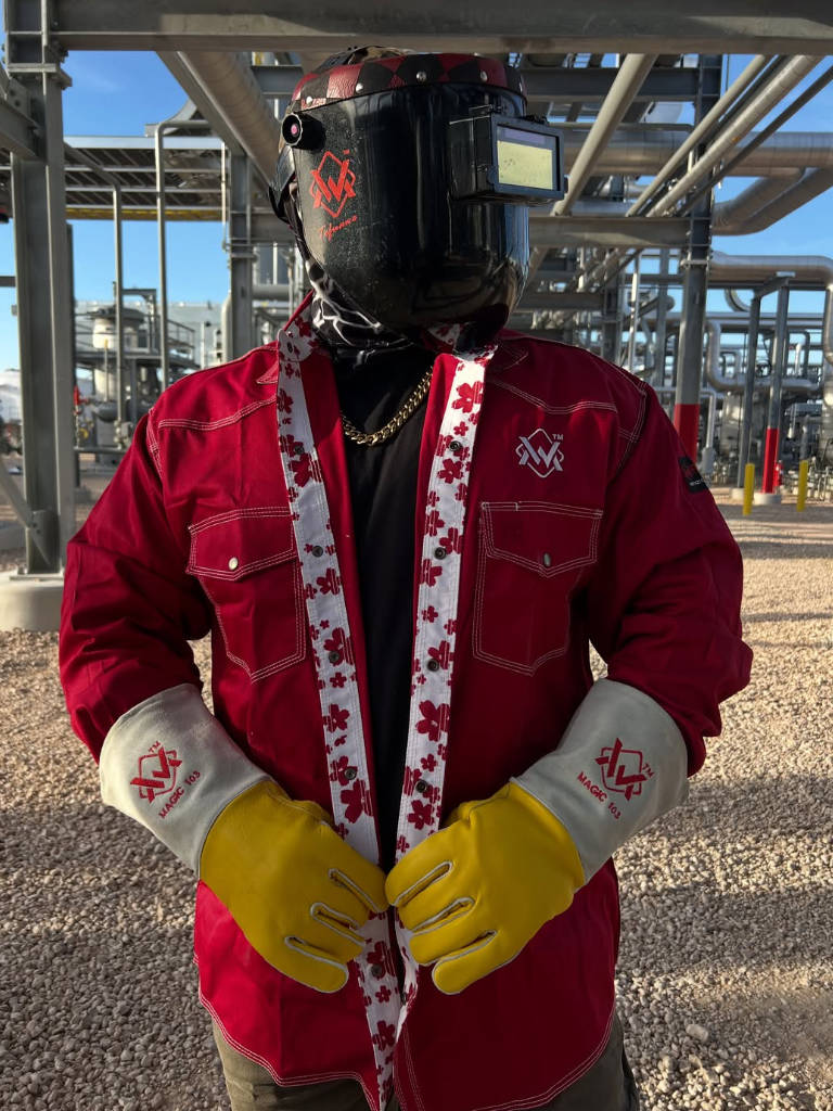 Person wearing a red jacket, yellow gloves, and a black welding helmet with a visible logo in an industrial setting.