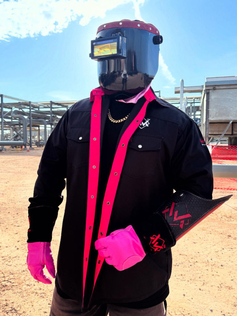 Person wearing a black shirt, pink gloves, and a welding helmet outdoors.
