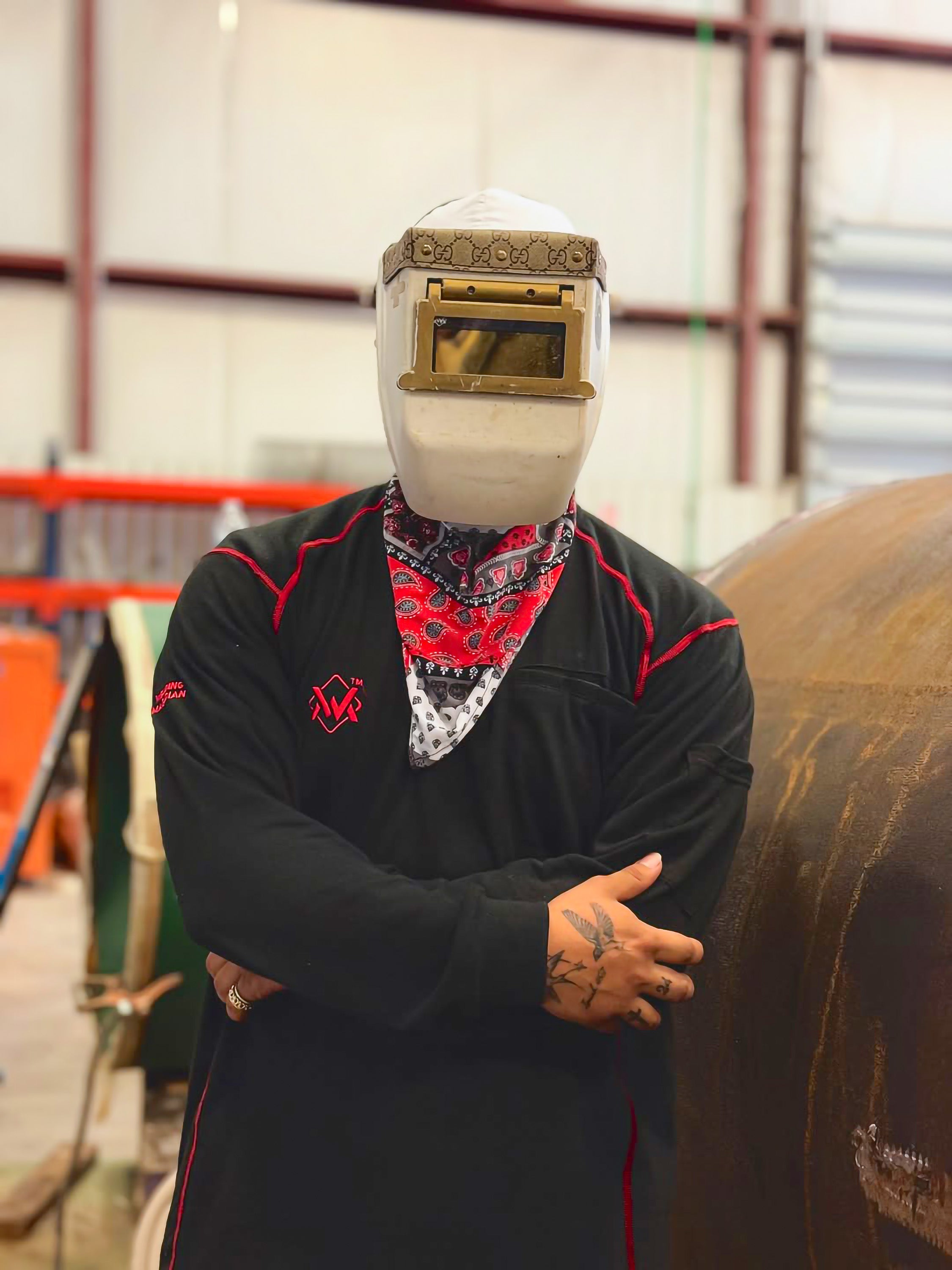 Person wearing a welding helmet and black jacket with red accents in an industrial setting