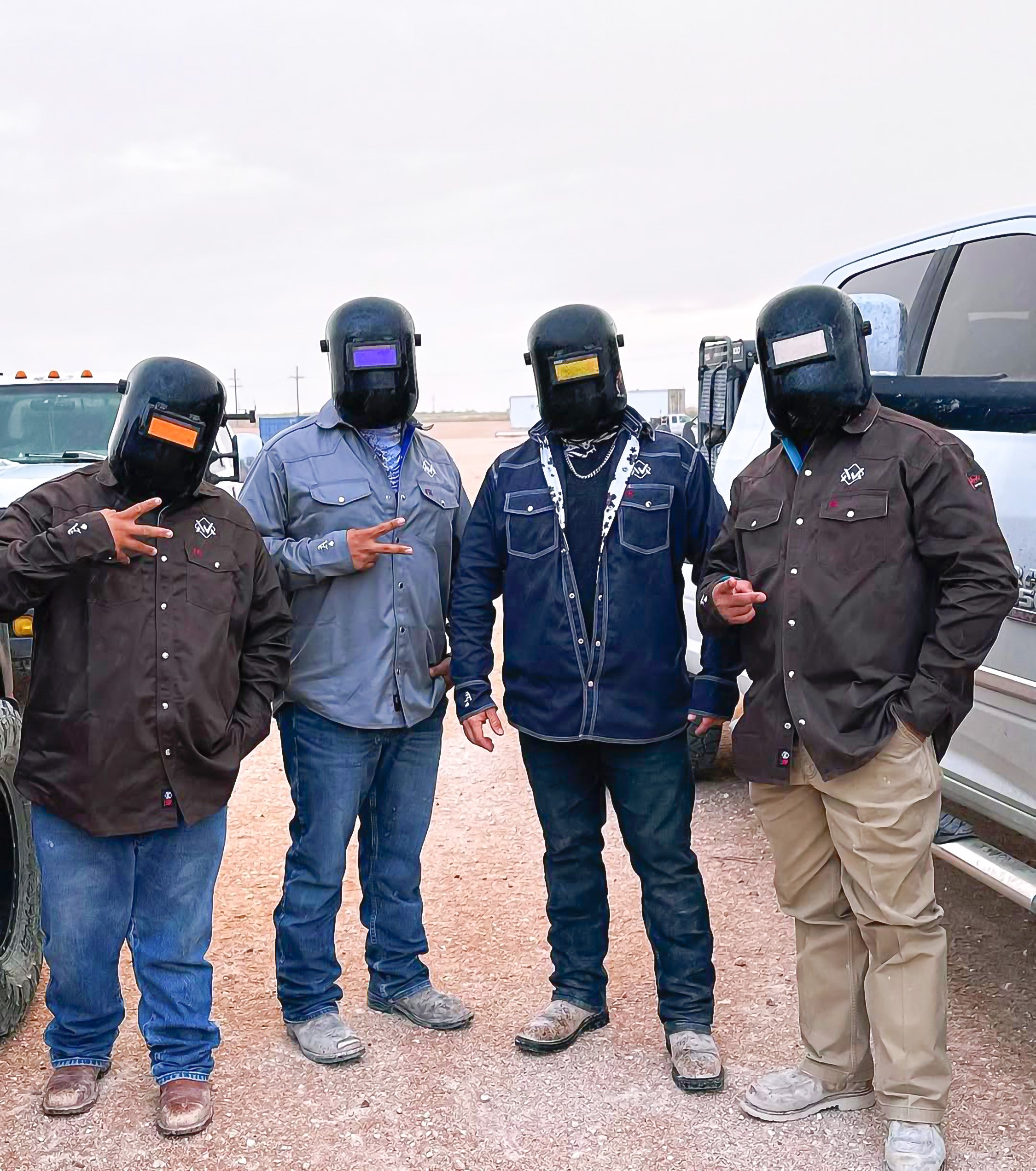 Four individuals wearing welding helmets and work attire standing outdoors.