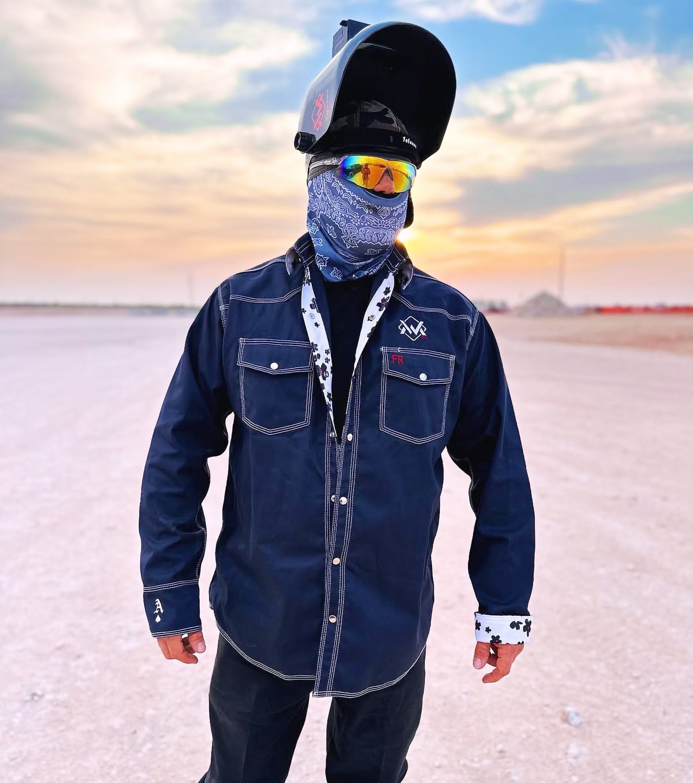 Person wearing a navy shirt and black cap with reflective goggles, standing on a salt flat at sunset.