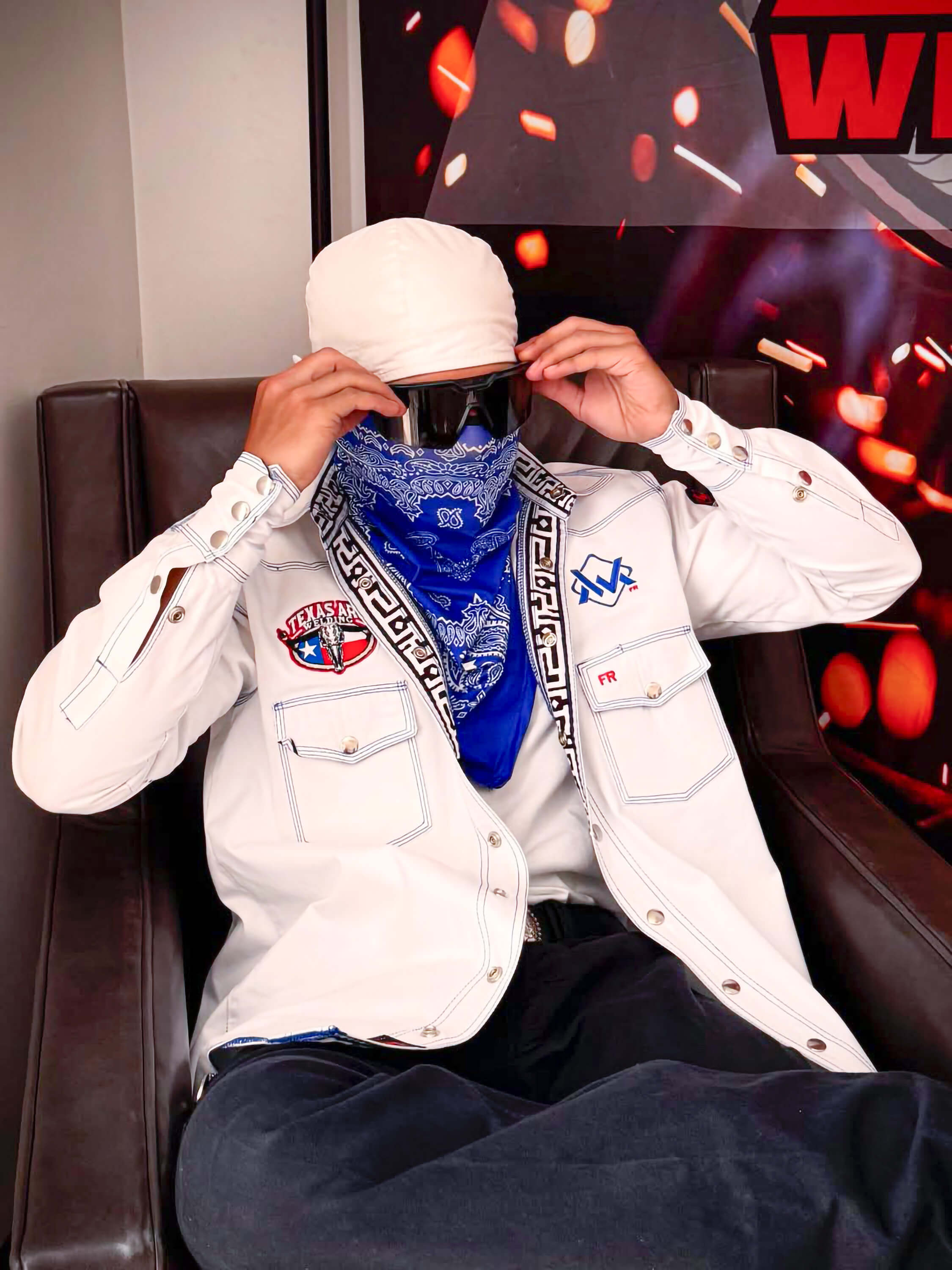 Person wearing a white jacket with logos, a blue bandana, and a white cap in front of a colorful background.