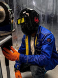 Person wearing a welding helmet and blue jacket with protective gloves, working on machinery.