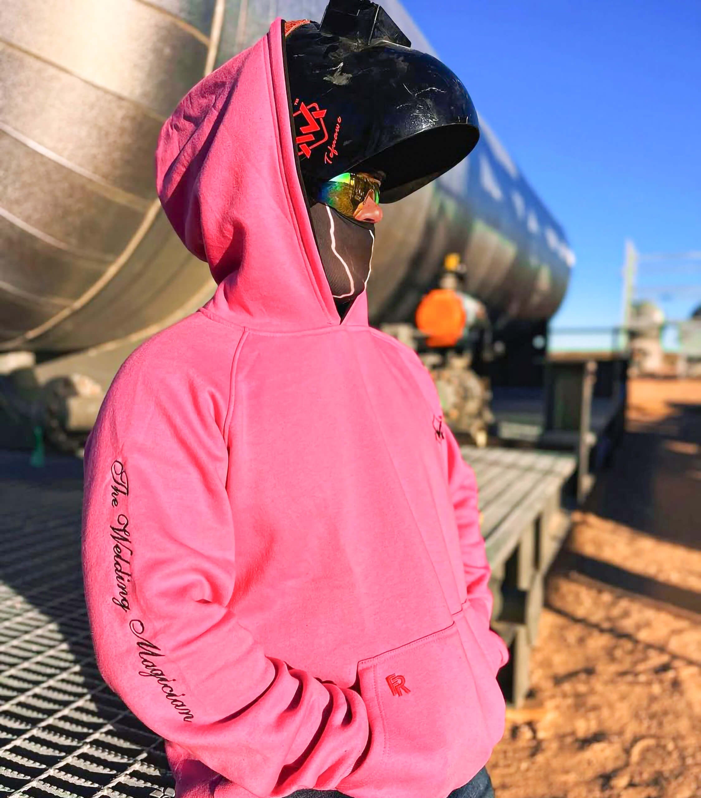 Person wearing a pink hoodie with a blurred face in front of large industrial tanks.