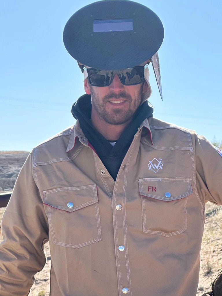 Man wearing a tan jacket with a logo, sunglasses, and a welding helmet outdoors.