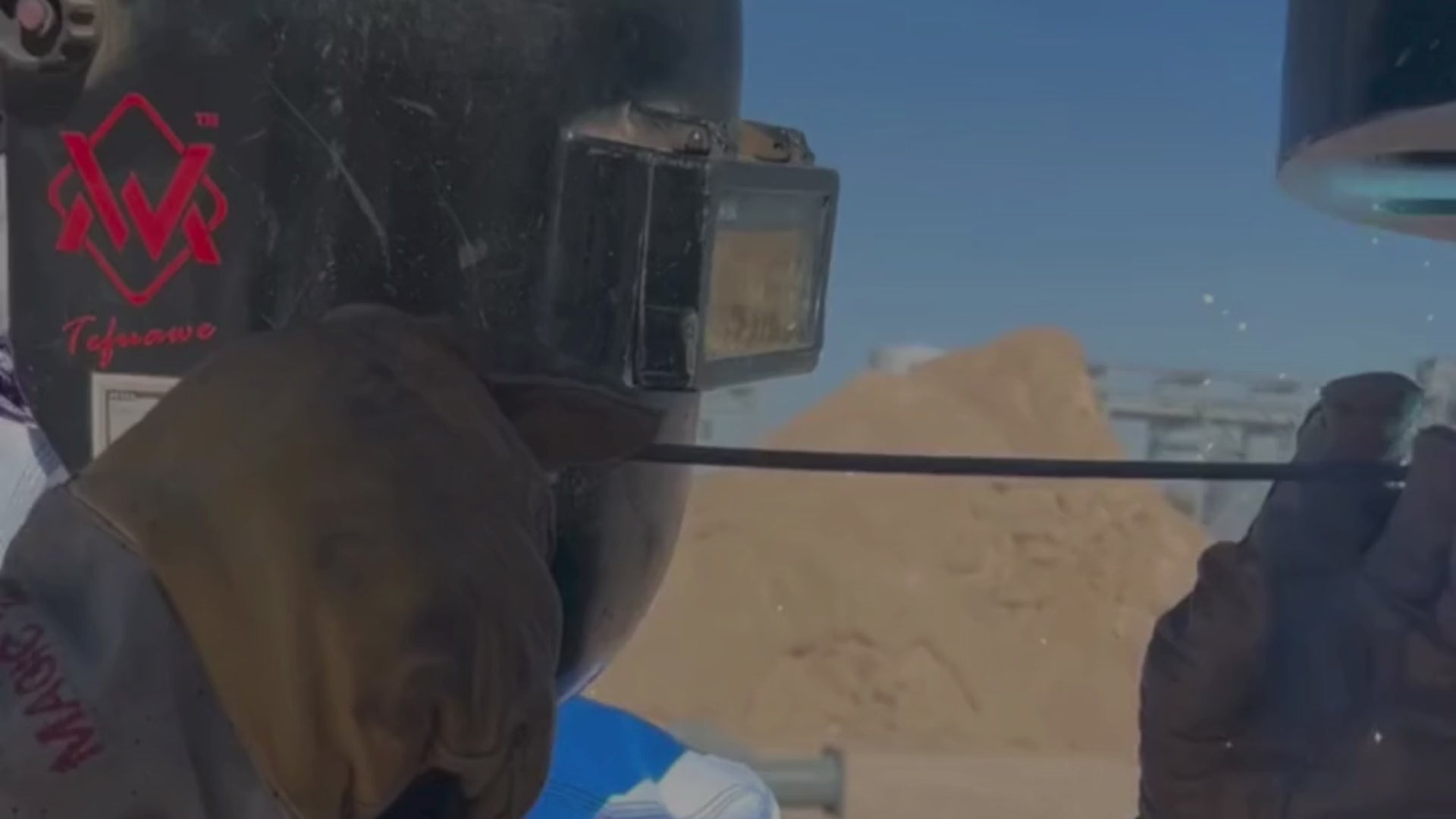 Load video: Welder performing field welding on a metal structure, wearing flame-resistant workwear under natural daylight conditions