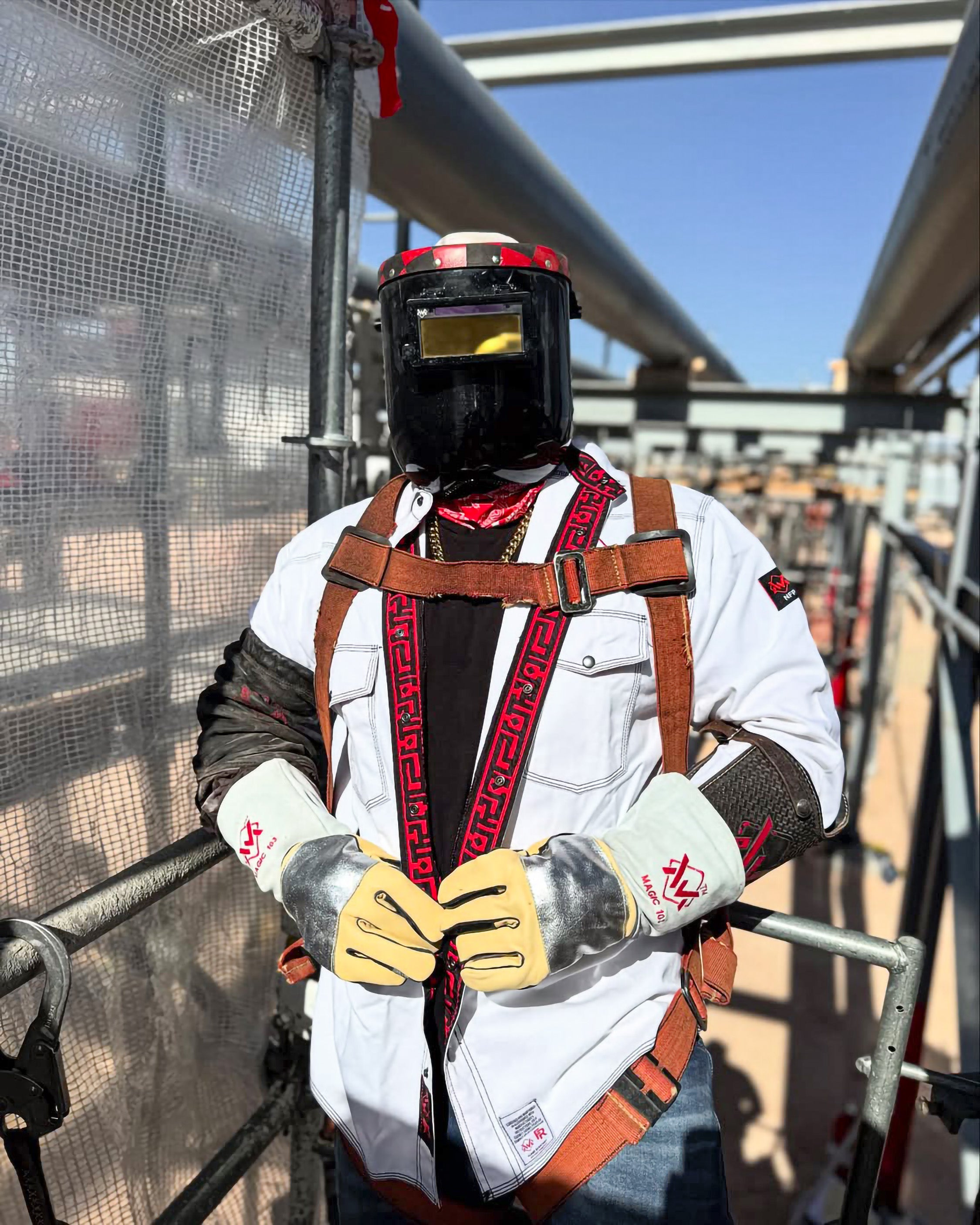 Person wearing a welding helmet, safety glasses, gloves, and a harness on a construction site.